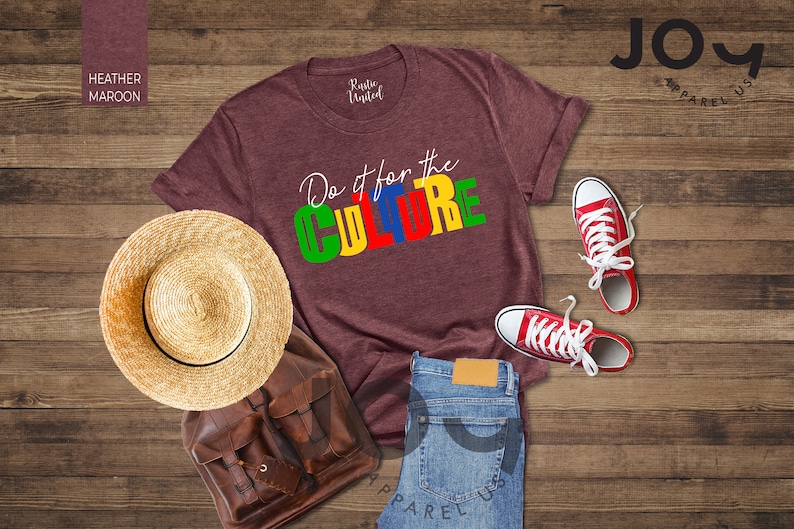 May include: Heather maroon t-shirt with the text "Do it for the CULTURE" in colorful block letters. The shirt is laid out on a wooden surface with a straw hat, a brown leather backpack, a pair of red sneakers, and blue jeans.
