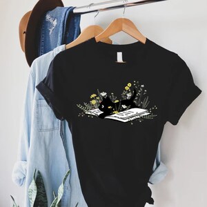 May include: Black t-shirt with a graphic of a black cat reading a book surrounded by flowers.