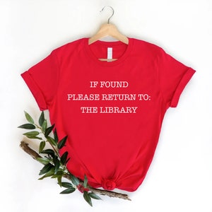 May include: A bright red short-sleeved t-shirt featuring white text printed on the front that reads, IF FOUND PLEASE RETURN TO: THE LIBRARY. The shirt is displayed on a wooden hanger, with a small branch of green leaves tucked underneath.