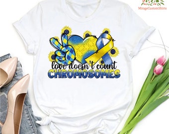 May include: White t-shirt with a blue and yellow peace sign, puzzle piece heart, and ribbon. The text "love doesn't count chromosomes" is printed below the design.