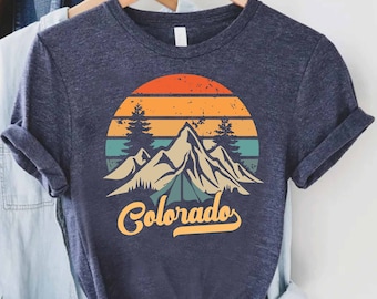 Puede incluir: Una camiseta azul oscuro con un gráfico de estilo vintage de montañas y una puesta de sol. El gráfico incluye el texto "Colorado" en una fuente retro.