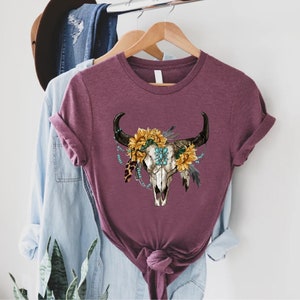 K&ouml;nnte beinhalten: Ein maroonfarbenes T-Shirt mit einem wei&szlig;en Totenkopf-Design. Der Totenkopf ist mit Sonnenblumen, t&uuml;rkisfarbenen Perlen und Federn verziert.