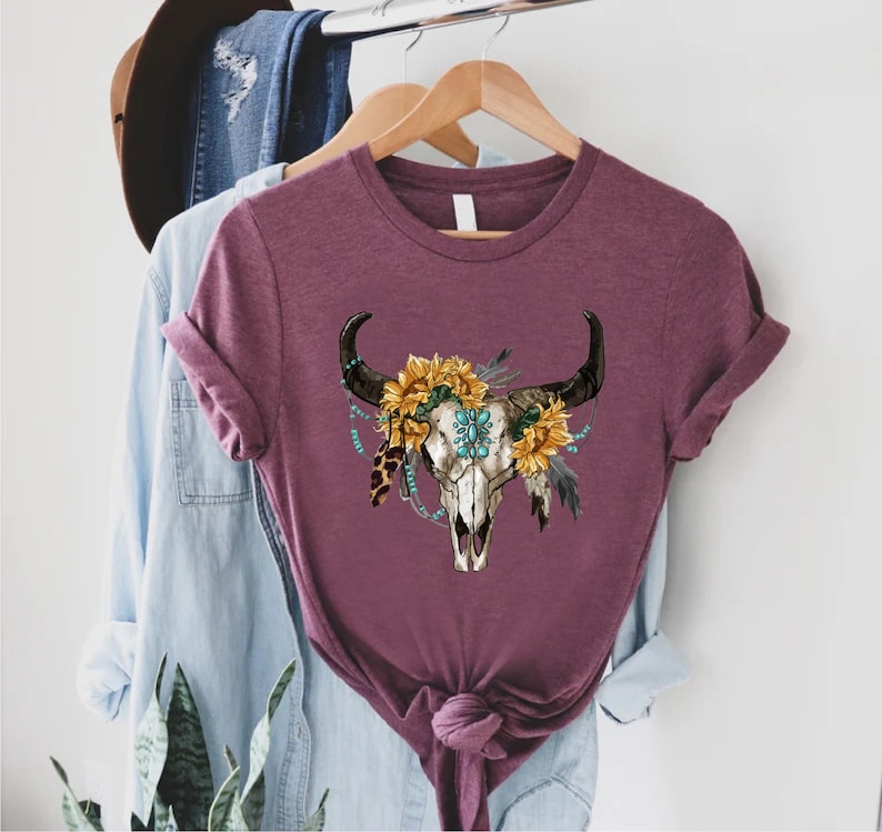 K&ouml;nnte beinhalten: Ein maroonfarbenes T-Shirt mit einem wei&szlig;en Totenkopf-Design. Der Totenkopf ist mit Sonnenblumen, t&uuml;rkisfarbenen Perlen und Federn verziert.