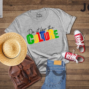 May include: A gray t-shirt with the text "Do it for the CULTURE" in colorful block letters. The shirt is laid out on a wooden surface with a straw hat, a brown leather backpack, a pair of red sneakers, and blue jeans.