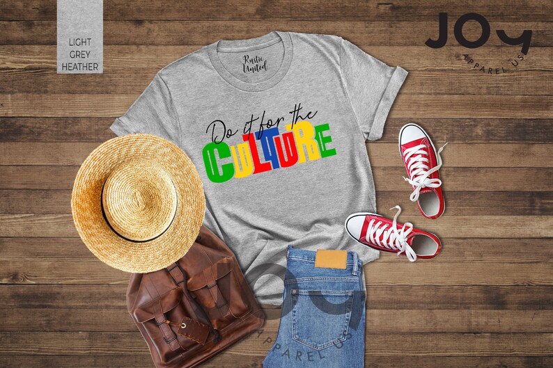 May include: A gray t-shirt with the text "Do it for the CULTURE" in colorful block letters. The shirt is laid out on a wooden surface with a straw hat, a brown leather backpack, a pair of red sneakers, and blue jeans.