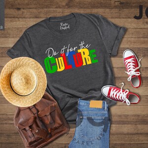 May include: A dark heather grey t-shirt with the text "Do it for the CULTURE" in colorful block letters. The shirt is laid out on a wooden surface with a straw hat, a brown leather backpack, and red sneakers.