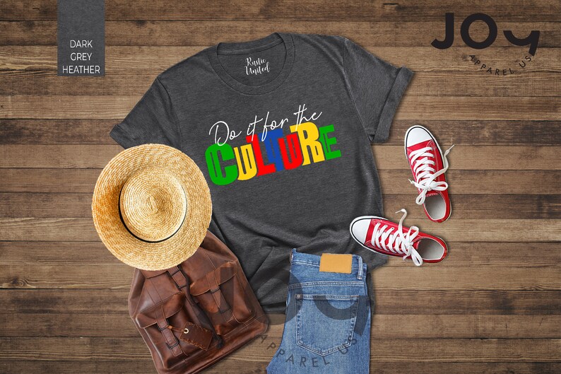 May include: A dark heather grey t-shirt with the text "Do it for the CULTURE" in colorful block letters. The shirt is laid out on a wooden surface with a straw hat, a brown leather backpack, and red sneakers.