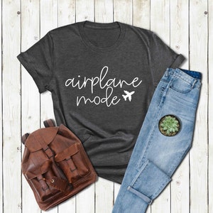 Puede incluir: Una camiseta gris oscuro con el texto "airplane mode" en letras blancas cursivas y un icono de avi&oacute;n blanco. La camiseta est&aacute; colocada sobre un fondo de madera blanca con una mochila de cuero marr&oacute;n y unos vaqueros azules.