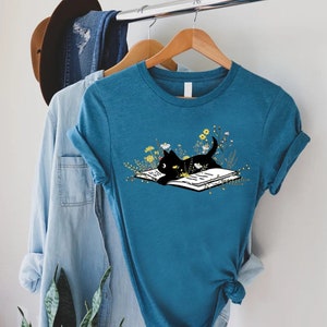May include: A teal blue t-shirt with a black cat reading a book surrounded by yellow flowers. The cat is lying on the book with its paws outstretched.