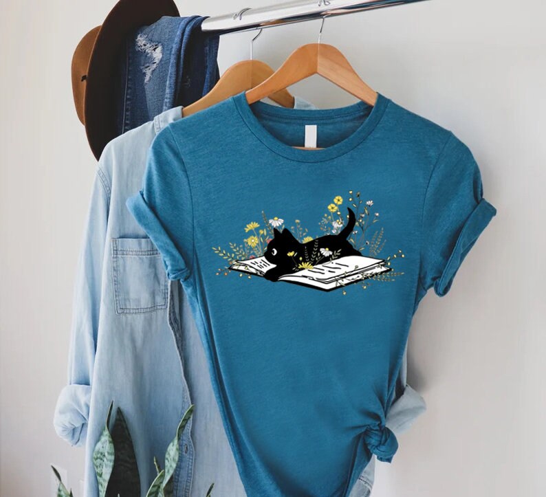 May include: A teal blue t-shirt with a black cat reading a book surrounded by yellow flowers. The cat is lying on the book with its paws outstretched.