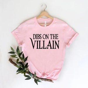 May include: Pink t-shirt with black text that reads "Dibs on the Villain".