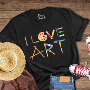 Puede incluir: Camiseta negra con las palabras "I LOVE ART" en letras de colores. Las letras est&aacute;n formadas por pinceles, l&aacute;pices y una paleta de pintura.