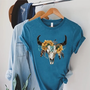 K&ouml;nnte beinhalten: Ein t&uuml;rkisfarbenes T-Shirt mit einem wei&szlig;en Totenkopf-Design. Der Totenkopf ist mit einer Sonnenblumenkrone, t&uuml;rkisfarbenen Perlen und Federn verziert.