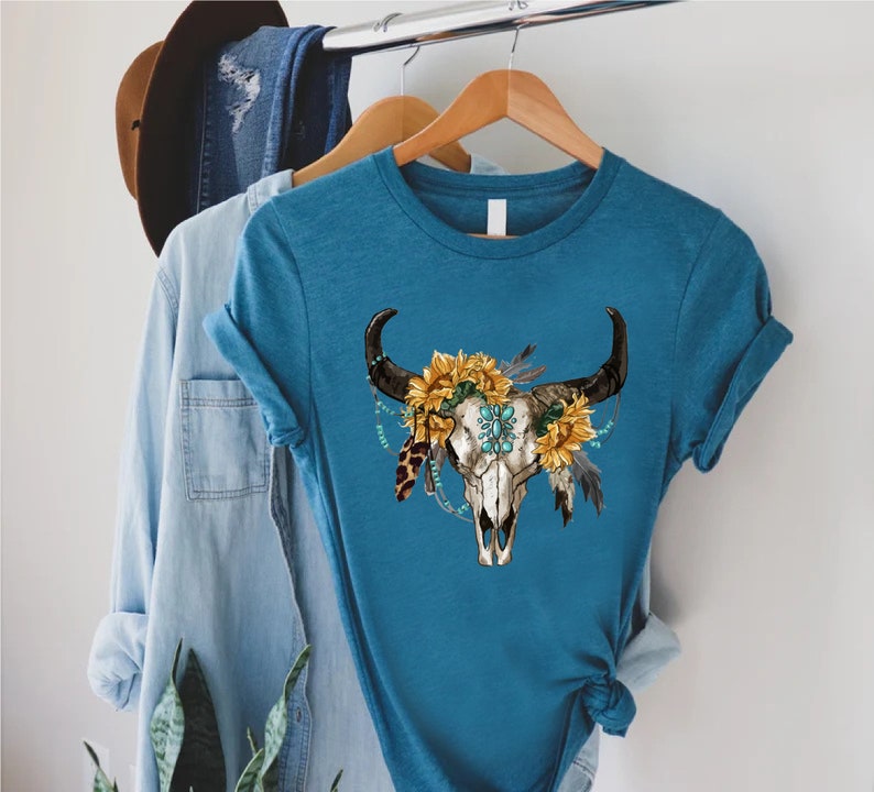 K&ouml;nnte beinhalten: Ein t&uuml;rkisfarbenes T-Shirt mit einem wei&szlig;en Totenkopf-Design. Der Totenkopf ist mit einer Sonnenblumenkrone, t&uuml;rkisfarbenen Perlen und Federn verziert.