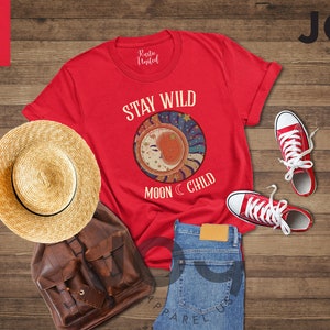 May include: Red t-shirt with the text "Stay Wild Moon Child" and a graphic of a crescent moon with stars. The shirt is laid out on a wooden surface with a straw hat, a brown leather backpack, blue jeans, and red trainers.