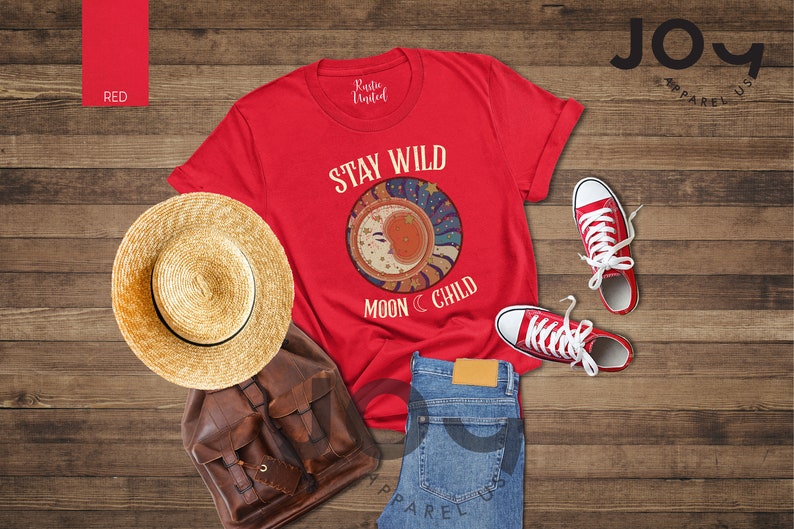 May include: Red t-shirt with the text "Stay Wild Moon Child" and a graphic of a crescent moon with stars. The shirt is laid out on a wooden surface with a straw hat, a brown leather backpack, blue jeans, and red trainers.