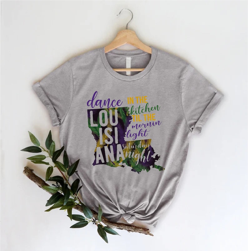 May include: Gray t-shirt with a purple and green outline of the state of Louisiana. The text "dance in the kitchen til the mornin light saturday night" is printed on the shirt.
