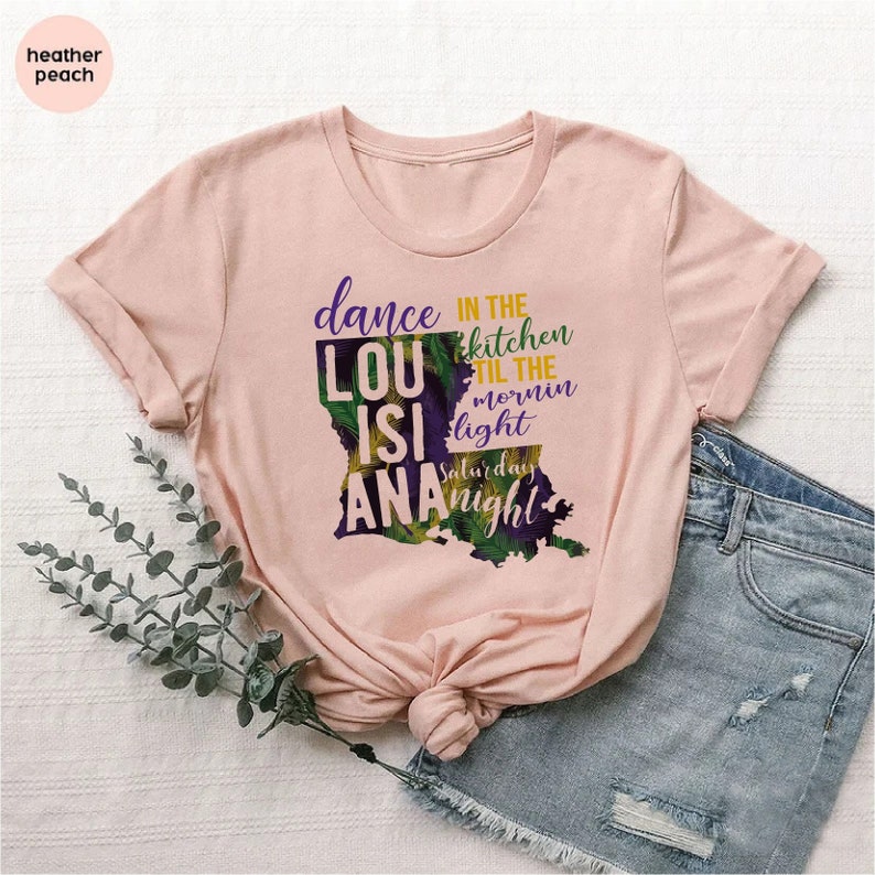 May include: A pink t-shirt with a graphic of the state of Louisiana. The graphic includes the text "dance in the kitchen til the mornin light Saturday night" and the words "LouisiAna".
