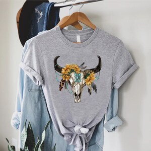 K&ouml;nnte beinhalten: Graues T-Shirt mit einem Aufdruck eines wei&szlig;en Bullensch&auml;dels, verziert mit Sonnenblumen, t&uuml;rkisfarbenen Perlen und Federn.