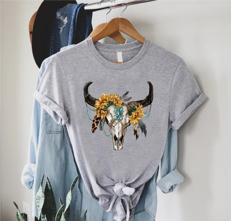 K&ouml;nnte beinhalten: Graues T-Shirt mit einem Aufdruck eines wei&szlig;en Bullensch&auml;dels, verziert mit Sonnenblumen, t&uuml;rkisfarbenen Perlen und Federn.