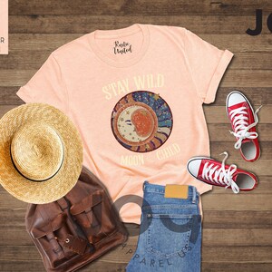 May include: A peach coloured t-shirt with the text "Stay Wild Moon Child" and a graphic of a crescent moon with stars and swirls. The shirt is laid out on a wooden surface with a straw hat, a brown leather backpack, and red trainers.