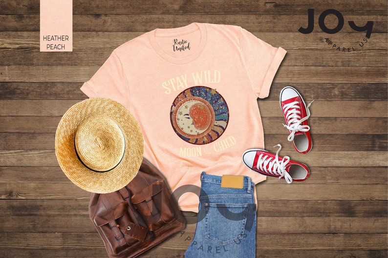 May include: A peach coloured t-shirt with the text "Stay Wild Moon Child" and a graphic of a crescent moon with stars and swirls. The shirt is laid out on a wooden surface with a straw hat, a brown leather backpack, and red trainers.