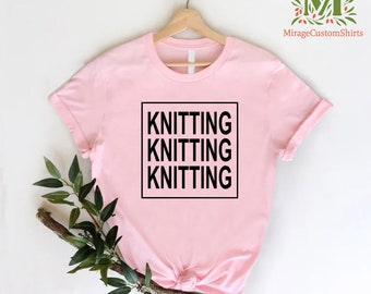 May include: A light pink t-shirt with the word "KNITTING" printed three times in a black, bold font inside a black square. The shirt is displayed on a wooden hanger against a white background. A sprig of greenery is placed on the lower left.