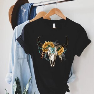 K&ouml;nnte beinhalten: Schwarzes T-Shirt mit einem wei&szlig;en Kuhsch&auml;del-Design. Der Sch&auml;del ist mit Sonnenblumen, t&uuml;rkisfarbenen Perlen und Federn verziert.