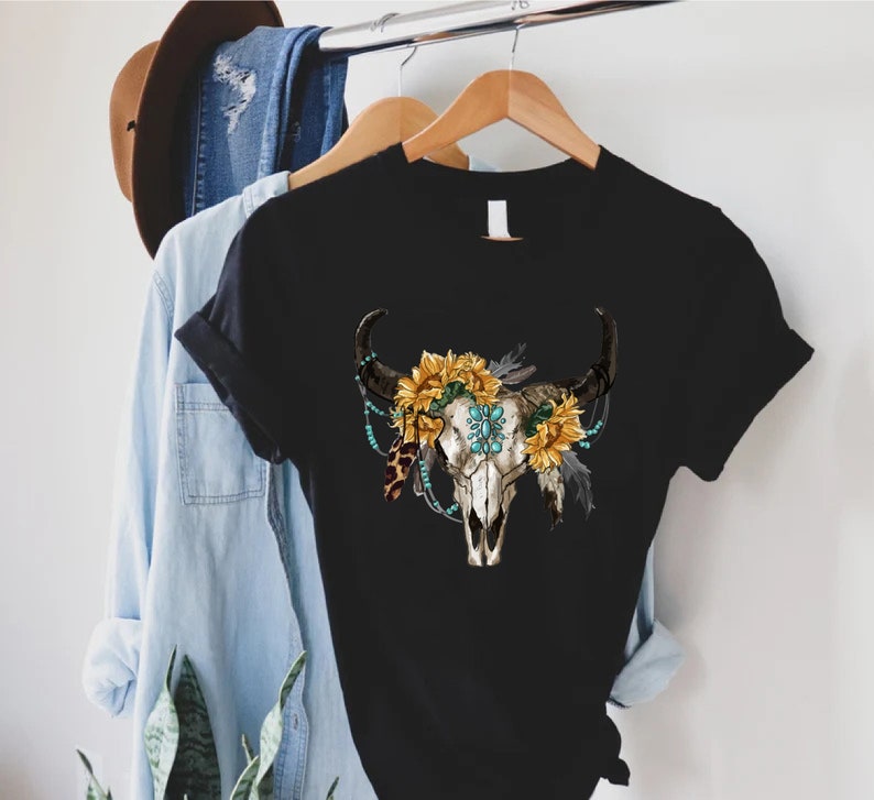 K&ouml;nnte beinhalten: Schwarzes T-Shirt mit einem wei&szlig;en Kuhsch&auml;del-Design. Der Sch&auml;del ist mit Sonnenblumen, t&uuml;rkisfarbenen Perlen und Federn verziert.
