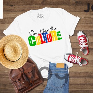 May include: White t-shirt with the text "Do it for the CULTURE" in colorful block letters. The shirt is laid out on a wooden surface with a straw hat, a brown leather bag, and red sneakers.
