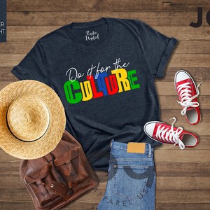 May include: A heather midnight navy t-shirt with the text "Do it for the CULTURE" in colorful block letters. The shirt is laid out on a wooden surface with a straw hat, a brown leather backpack, and red sneakers.