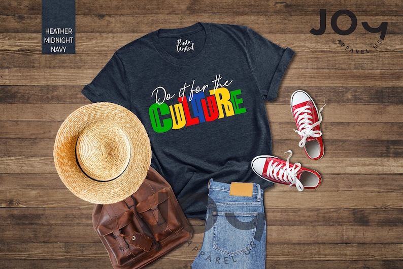 May include: A heather midnight navy t-shirt with the text "Do it for the CULTURE" in colorful block letters. The shirt is laid out on a wooden surface with a straw hat, a brown leather backpack, and red sneakers.