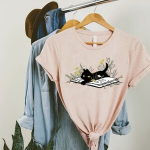 May include: A pink t-shirt with a black cat reading a book surrounded by flowers. The cat is looking at the book.