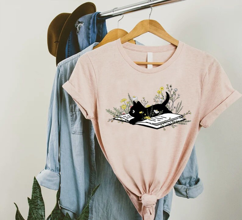 May include: A pink t-shirt with a black cat reading a book surrounded by flowers. The cat is looking at the book.