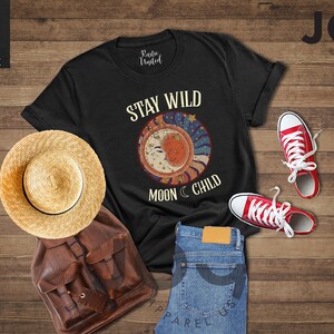 May include: Black t-shirt featuring a graphic of a crescent moon and stars with the text "Stay Wild Moon Child". The shirt is laid out on a wooden surface with a straw hat, a brown leather backpack, and red trainers.