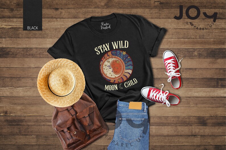 May include: Black t-shirt featuring a graphic of a crescent moon and stars with the text "Stay Wild Moon Child". The shirt is laid out on a wooden surface with a straw hat, a brown leather backpack, and red trainers.