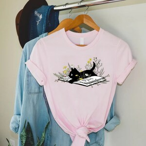 May include: A pink t-shirt with a black cat reading a book surrounded by flowers. The cat is lying on the book with its head resting on the page. The flowers are yellow and white.