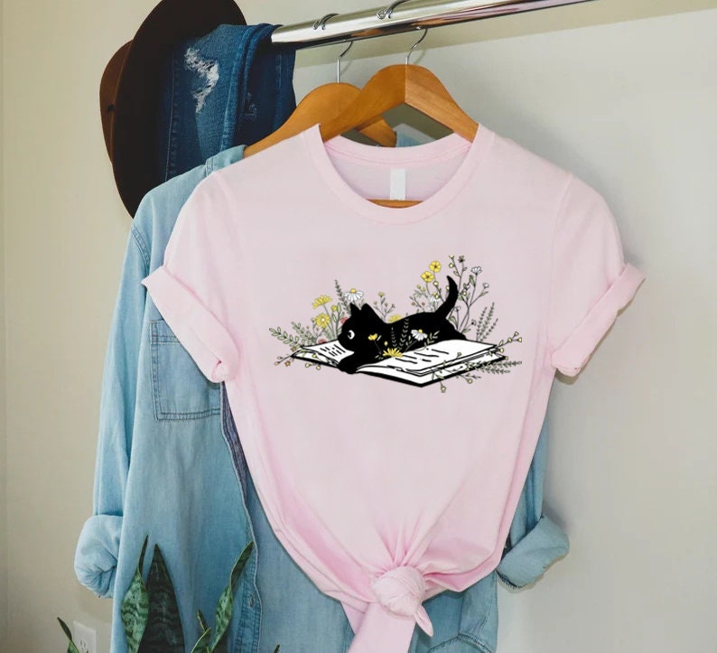 May include: A pink t-shirt with a black cat reading a book surrounded by flowers. The cat is lying on the book with its head resting on the page. The flowers are yellow and white.