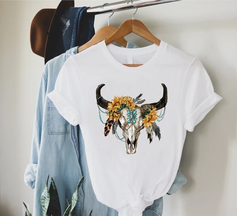 K&ouml;nnte beinhalten: Wei&szlig;es T-Shirt mit einem Aufdruck eines Kuhsch&auml;dels mit einer Sonnenblumenkrone und t&uuml;rkisfarbenen Perlen.