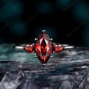 Può includere: Un anello con una pietra preziosa rossa centrale taglio marquise, affiancata da due pietre preziose rosse più piccole taglio a goccia su una fascia in metallo scuro. L'anello è fissato con griffe che tengono le pietre saldamente in posizione.