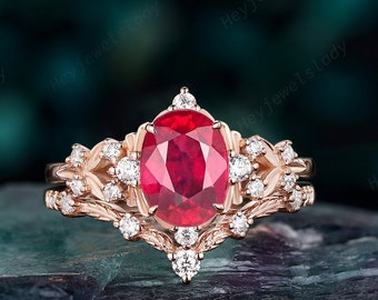 May include: A rose gold ring set featuring a central oval-cut red gemstone surrounded by small round white stones. The band and setting are crafted with intricate leaf-like metalwork details, creating a layered appearance with a matching band.