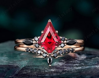 May include: A rose gold ring set featuring a large, kite-shaped red gemstone as the centerpiece. The ring is accented with black gemstones and a matching band. The design is intricate and elegant, suitable for special occasions.