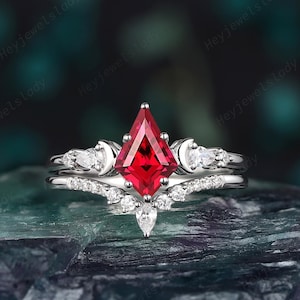 Puede incluir: Un conjunto de anillos en tono plateado con una piedra central roja en forma de cometa, flanqueada por dos detalles de media luna y pequeñas piedras claras. Incluye una banda a juego en forma de chevrón adornada con piedras claras.