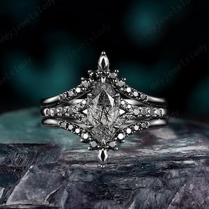 May include: A black metal ring set featuring a central marquise-cut rutilated quartz stone with black needle-like inclusions. The band is adorned with small black stones in a symmetrical, pointed design that extends above and below the center.