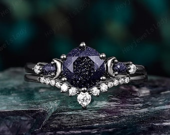 May include: A black gold engagement ring set with a large blue gemstone and smaller accent stones. The center stone is a square cut with a textured surface. The band is a delicate design with small round stones.