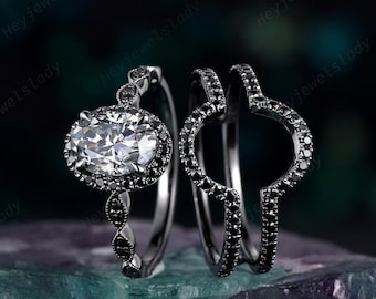 May include: A set of three rings. The engagement ring features a large, oval-cut, clear gemstone surrounded by black stones. The band has a delicate design. The other two rings are black and curved, designed to fit around the engagement ring.