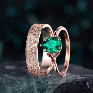 Könnte beinhalten: Ein Paar Trauringe aus Roségold mit einem keltischen Knotendesign. Der größere Ring hat einen birnenförmigen smaragdgrünen Edelstein.