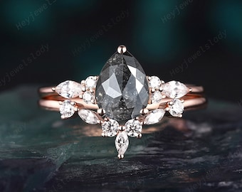 Puede incluir: Un anillo de oro rosa con un diamante central en forma de pera tipo sal y pimienta. La banda está decorada con diamantes blancos más pequeños de talla marquesa y redonda, dispuestos en un diseño de racimo que enmarca la piedra principal.