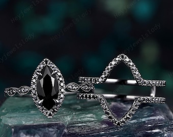 May include: A black metal ring set featuring a marquise-cut black center stone surrounded by a halo of small black stones, paired with a matching chevron-shaped band encrusted with tiny black stones along the entire outer edge of the design.