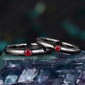 Può includere: Due fedi nuziali nere con un singolo gioiello rosso su ogni anello. Gli anelli sono impilati uno sopra l'altro.